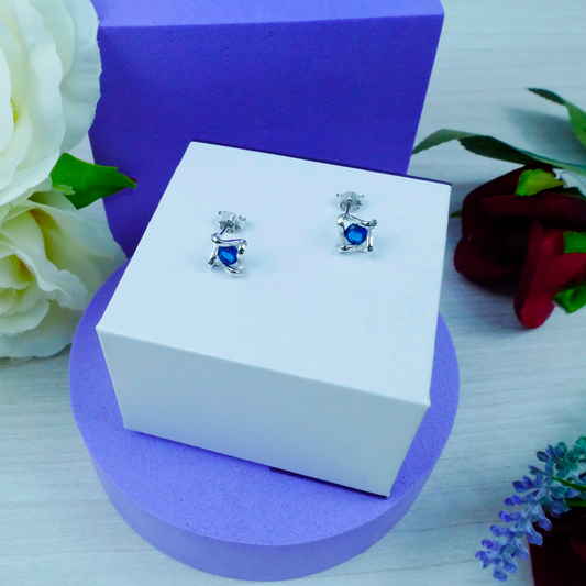 Silver earrings with blue gemstones on a white box with a purple base, surrounded by flowers.