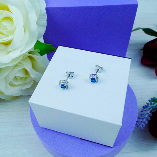 Silver earrings with blue stones on a white box with purple base, surrounded by flowers.
