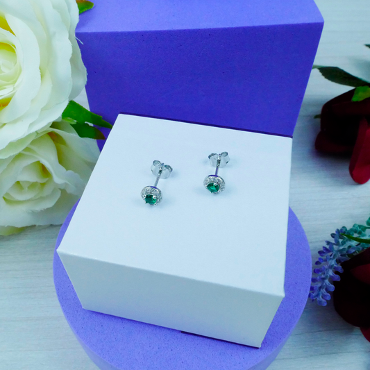 Silver earrings with green gemstones on a white box with a purple base, surrounded by flowers.