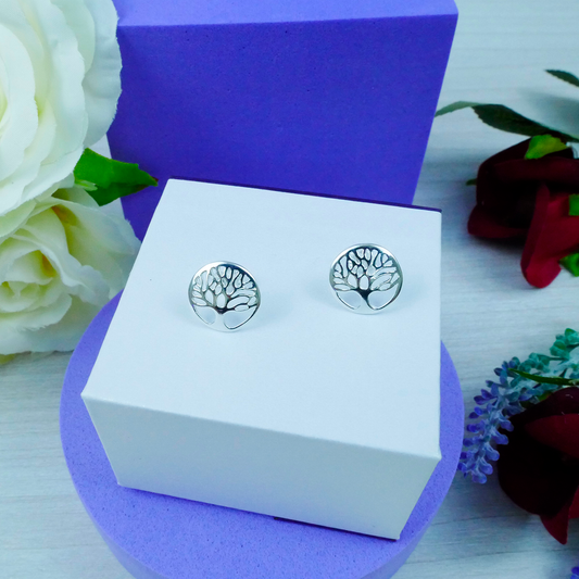 Silver tree-shaped earrings on a white box with a purple base, surrounded by flowers.