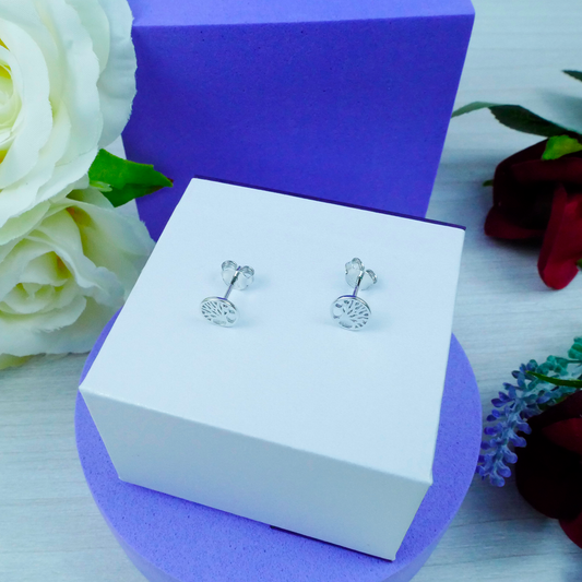 Silver tree-shaped earrings on a white box with purple base, surrounded by flowers.