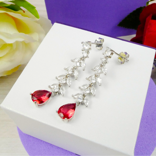 Silver earrings with red gemstones on a white box with purple accents, surrounded by flowers.