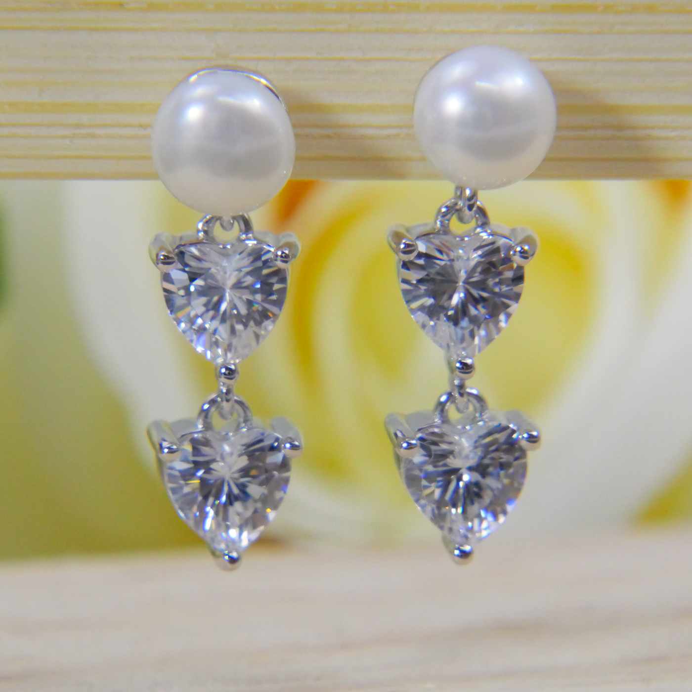 Pearl and heart-shaped Natrual Zircon gemstone earrings on a wooden surface with a blurred floral background.