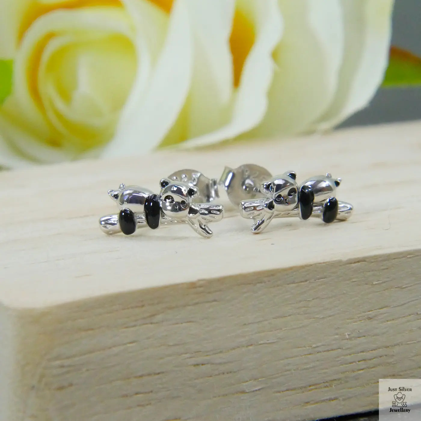Silver panda-shaped earrings on a wooden surface with a blurred yellow rose in the background
