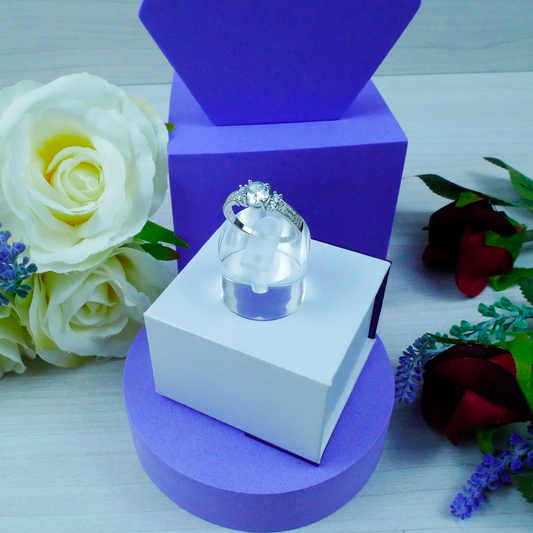 Silver ring on a white box with purple base, surrounded by flowers on a light wooden surface