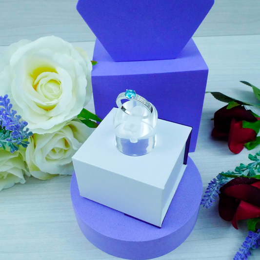 Ring on a white box with purple base, surrounded by flowers on a light blue background