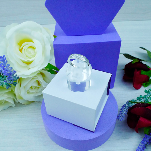 Silver ring in a clear glass dome on a white box with purple base, surrounded by flowers.