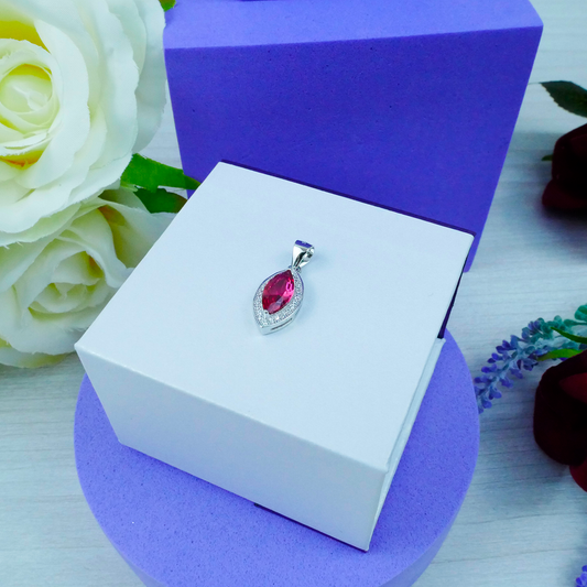 Jewelry pendant with a red gemstone in a white box on a purple stand, with flowers in the background.