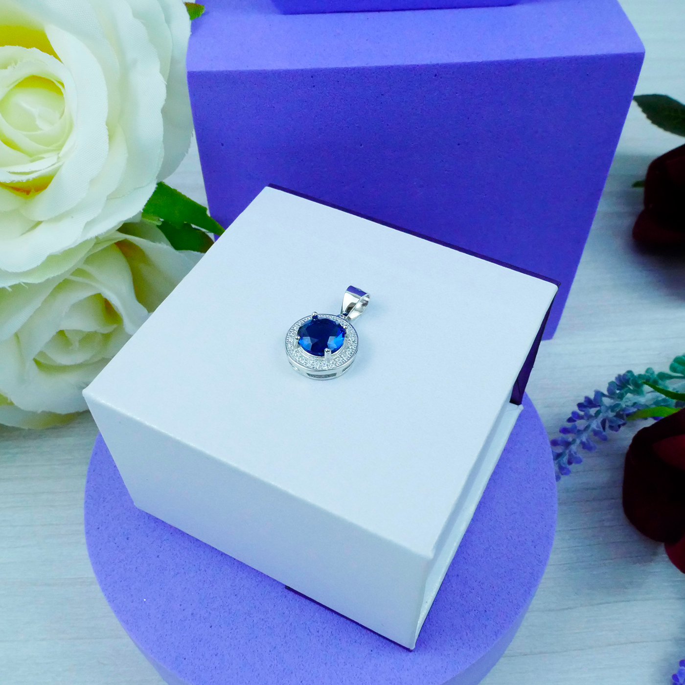 Jewelry pendant with blue gemstone on a white box with purple base, surrounded by flowers.