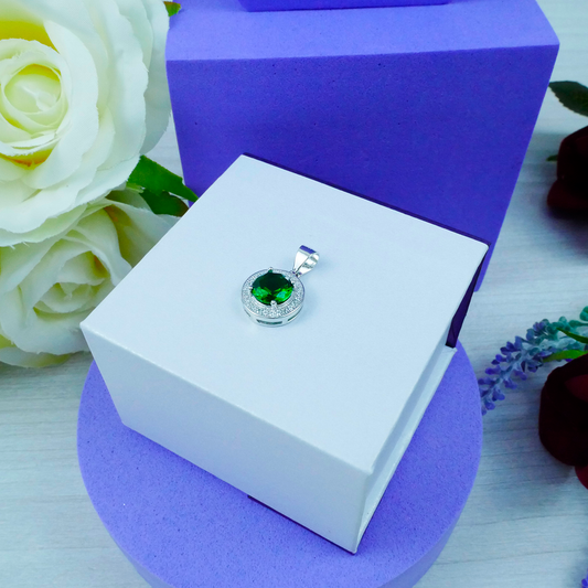 Silver pendant with a green gemstone on a white box with purple base, surrounded by flowers.