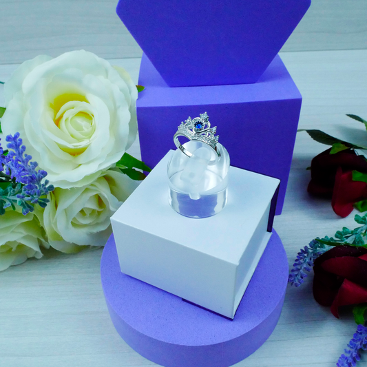 Clear glass heart with silver crown on a white and purple stand, surrounded by flowers.
