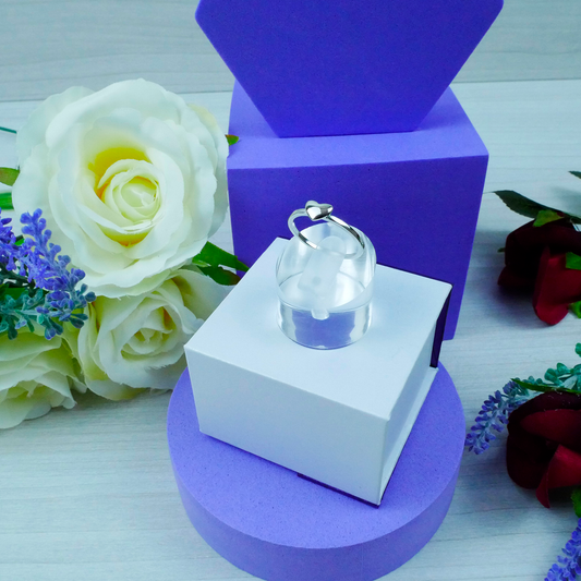 Silver ring in a clear glass box on a white and purple stand with flowers in the background