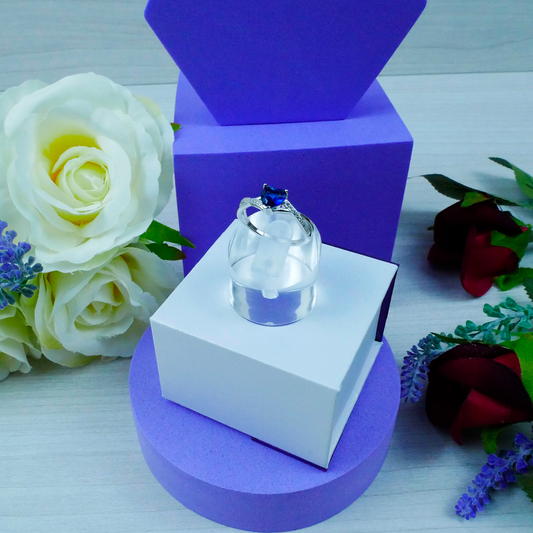 Clear crystal heart with blue stone on a white and purple stand, surrounded by flowers.