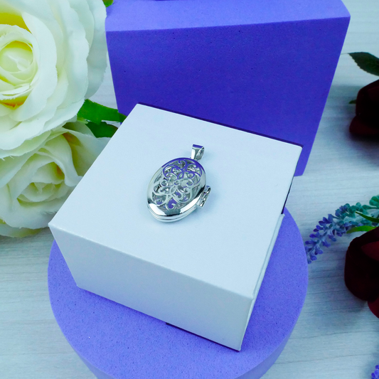 Silver locket in a white box with purple interior, surrounded by flowers.