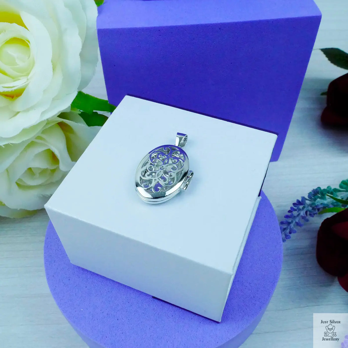 Silver locket in a white box with purple interior, surrounded by flowers.