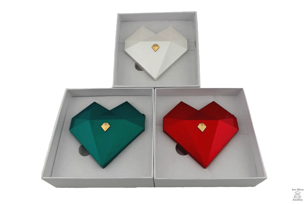 Three heart-shaped ring gift boxes in white, green, and red, with gold accents, displayed in an open presentation box.