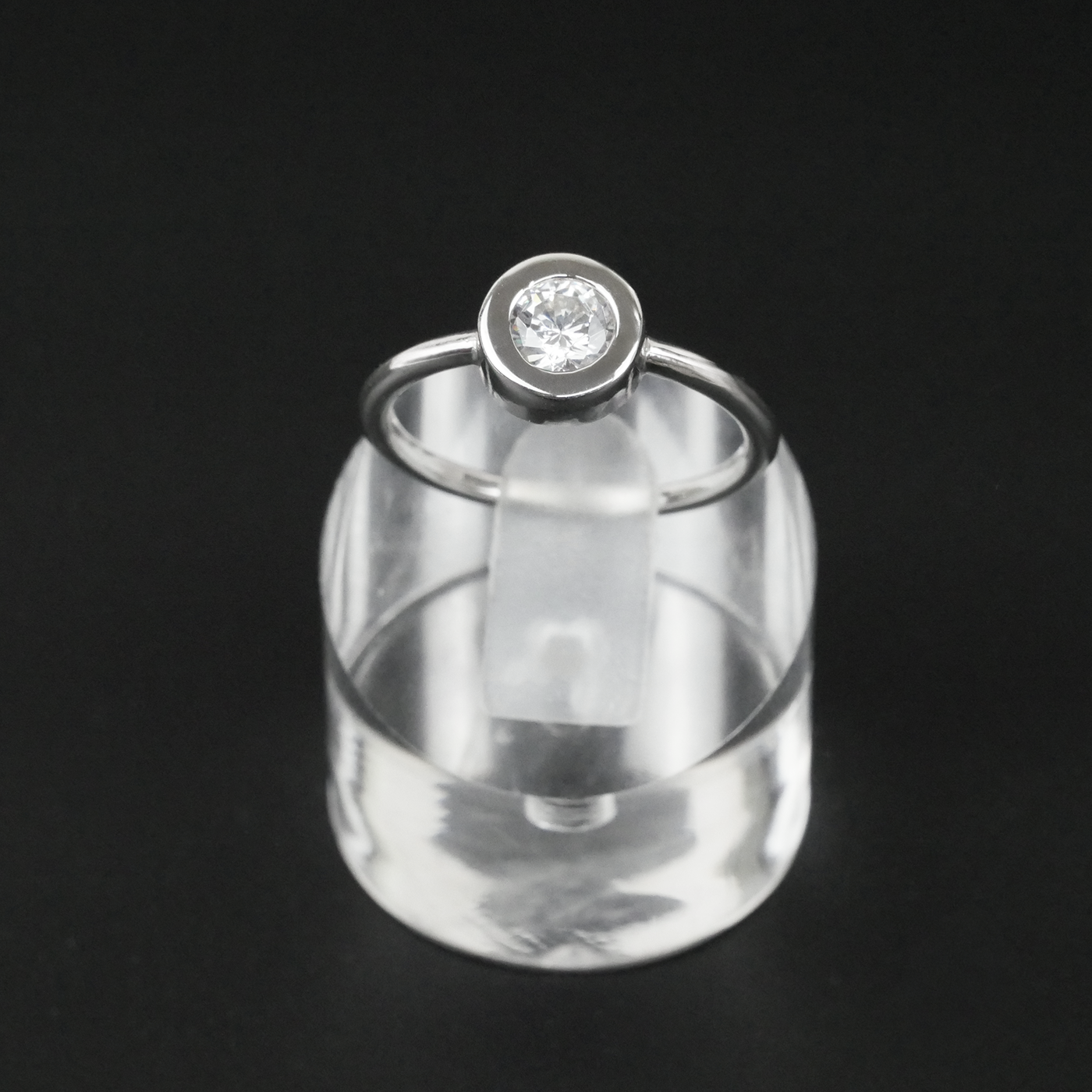 Silver ring with a CZ on a clear cylindrical stand against a black background