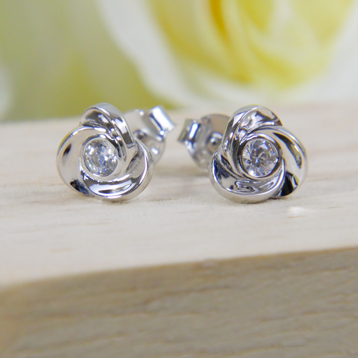 Silver earrings with a central zircon gemstone on a wooden surface with a blurred yellow flower in the background