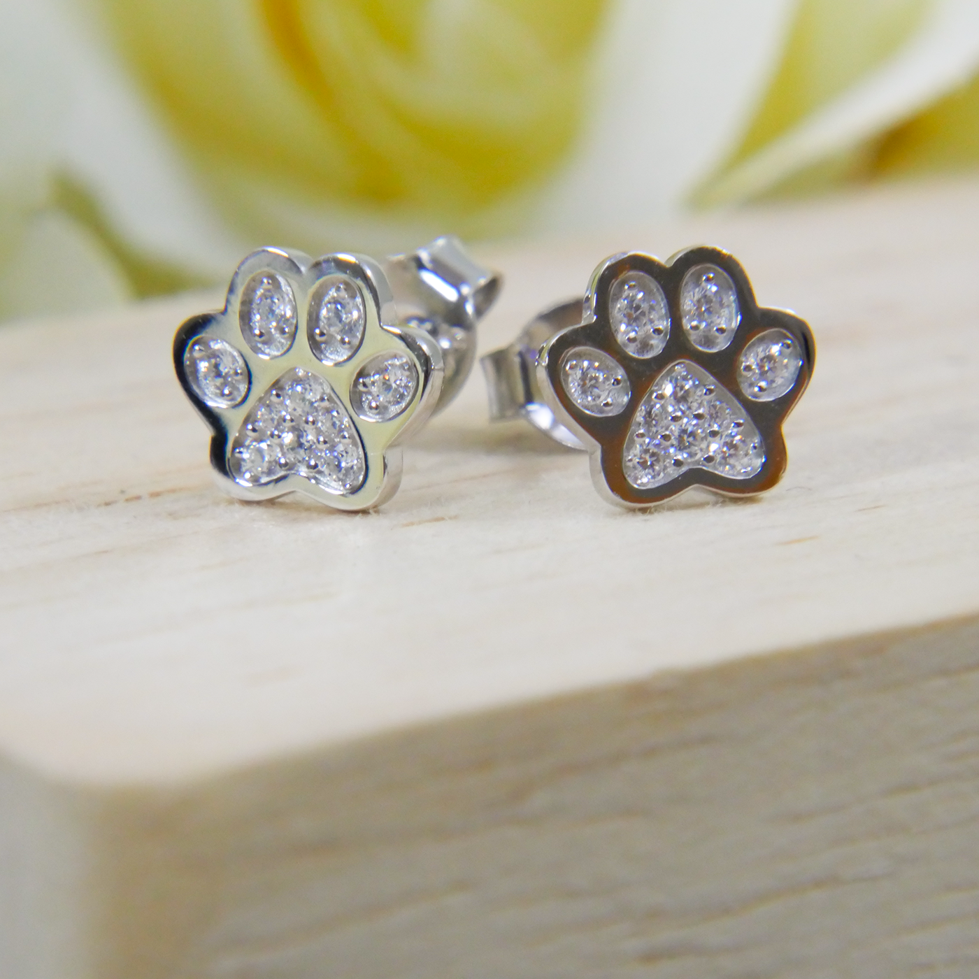 Paw print earrings with clear zircon stones on a wooden surface with a blurred floral background