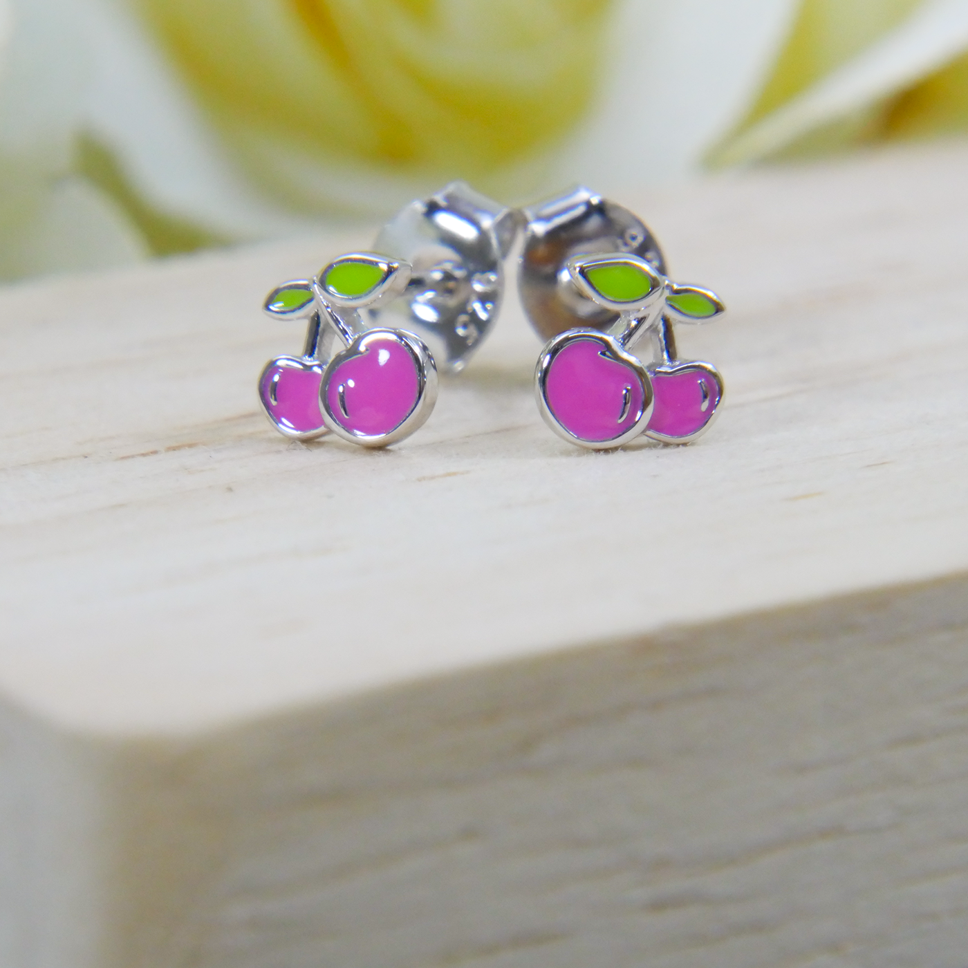 Cherry-themed stud earrings on a wooden surface with a blurred floral background