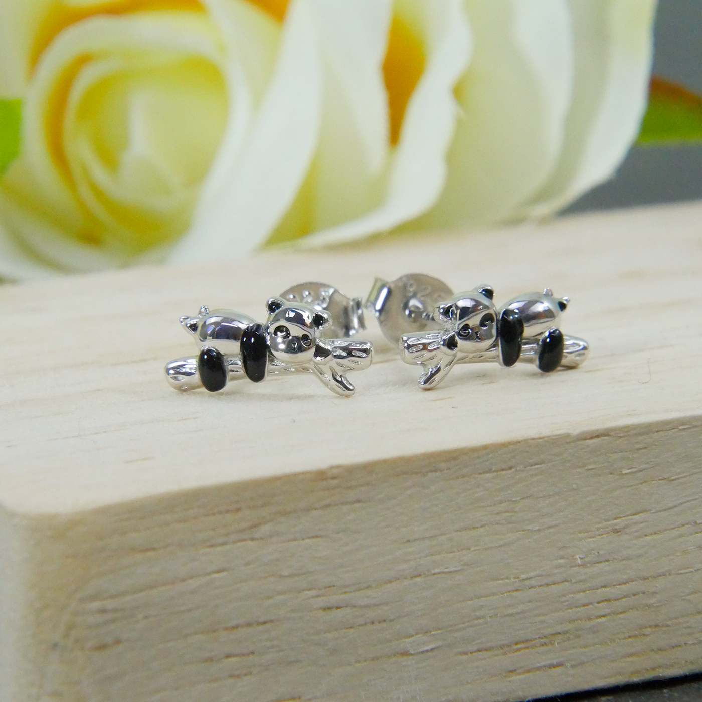 Silver panda-shaped earrings on a wooden surface with a blurred yellow rose in the background