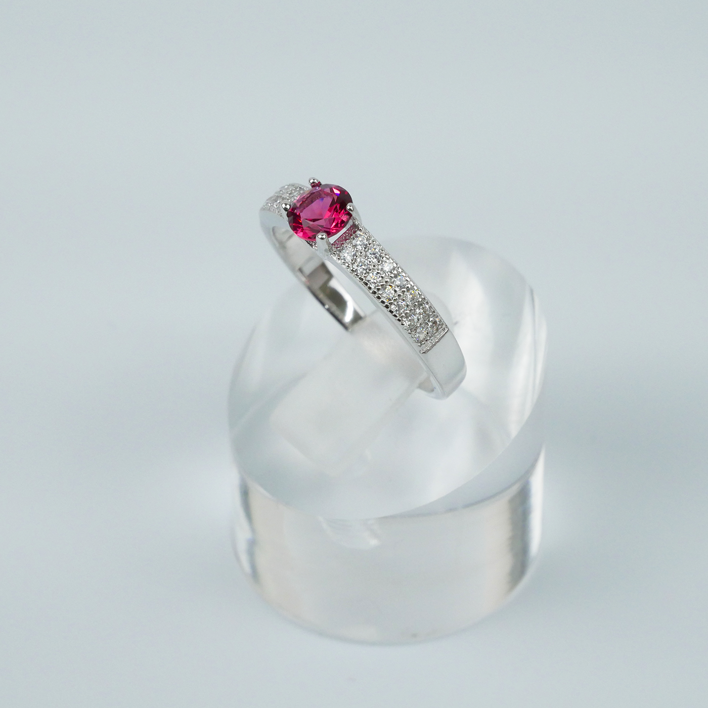 Silver ring with a red gemstone on a clear stand against a light-grey background