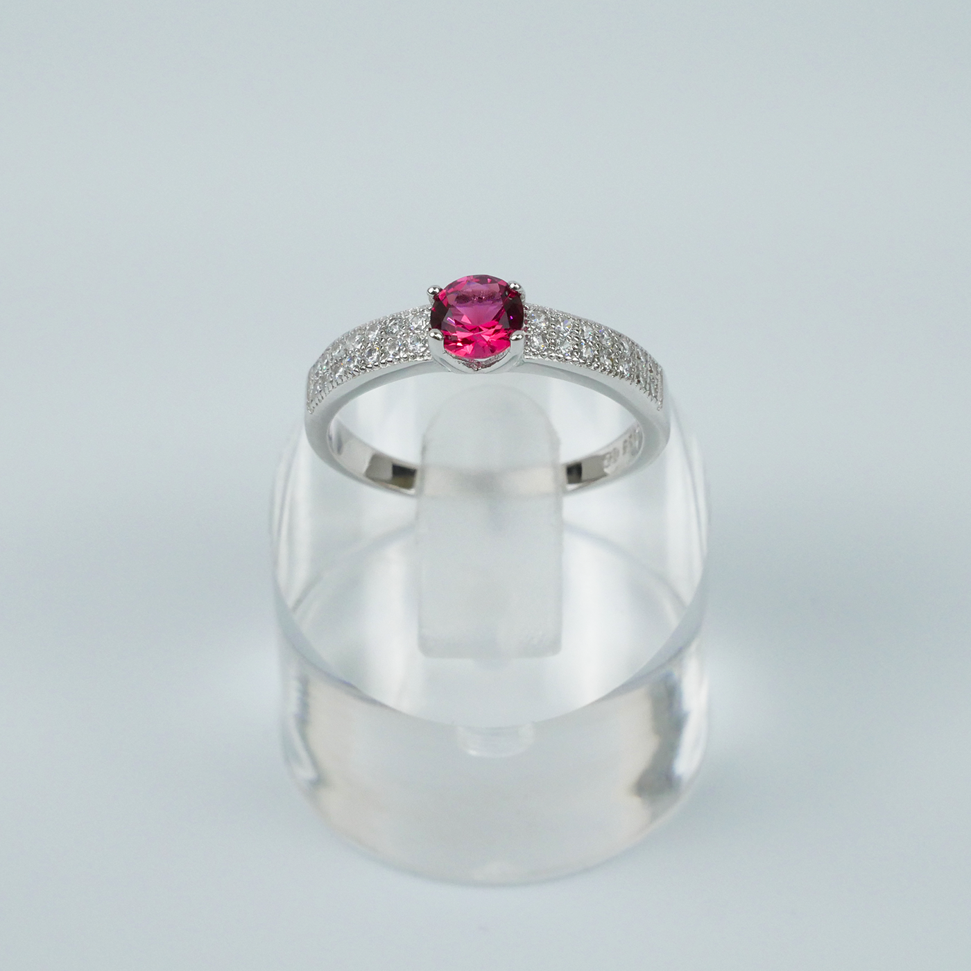Silver ring with a red gemstone on a clear stand against a light-grey background