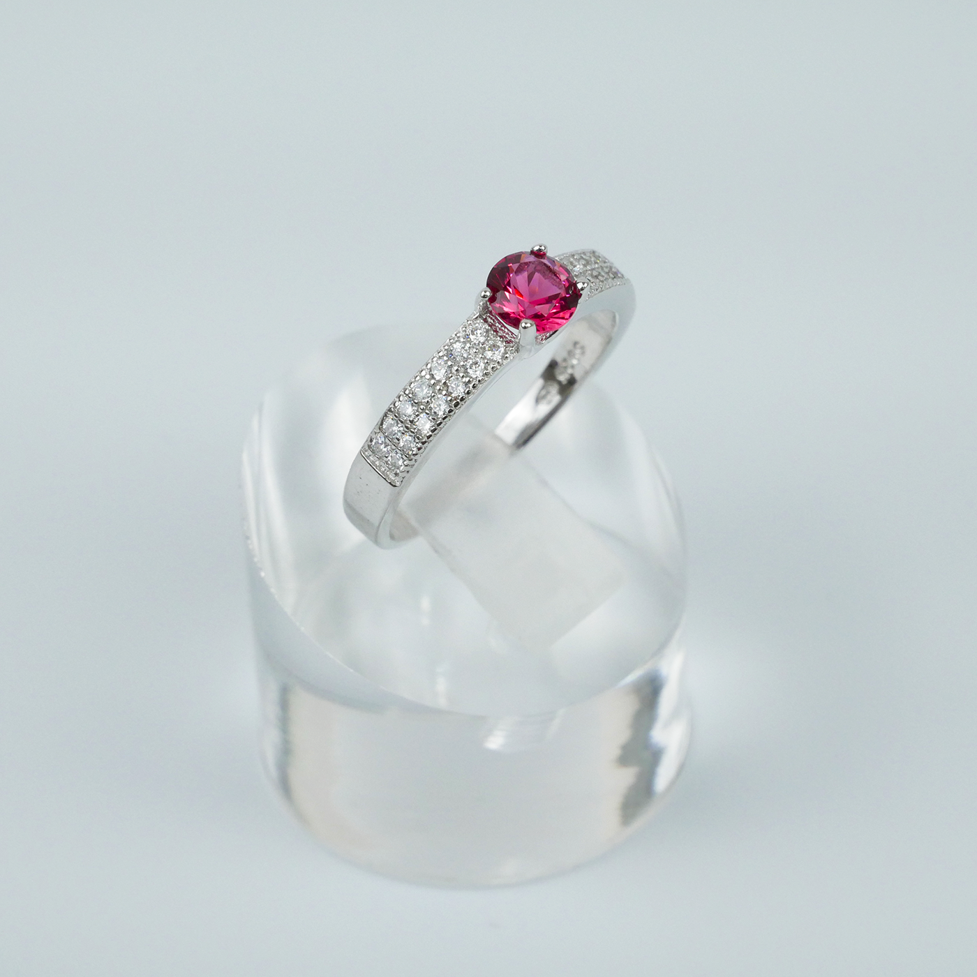 Silver ring with a red gemstone on a clear stand against a light-grey background