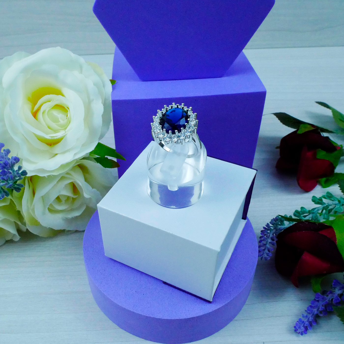 Jewelry box with a ring featuring a blue gemstone, surrounded by flowers on a white surface.