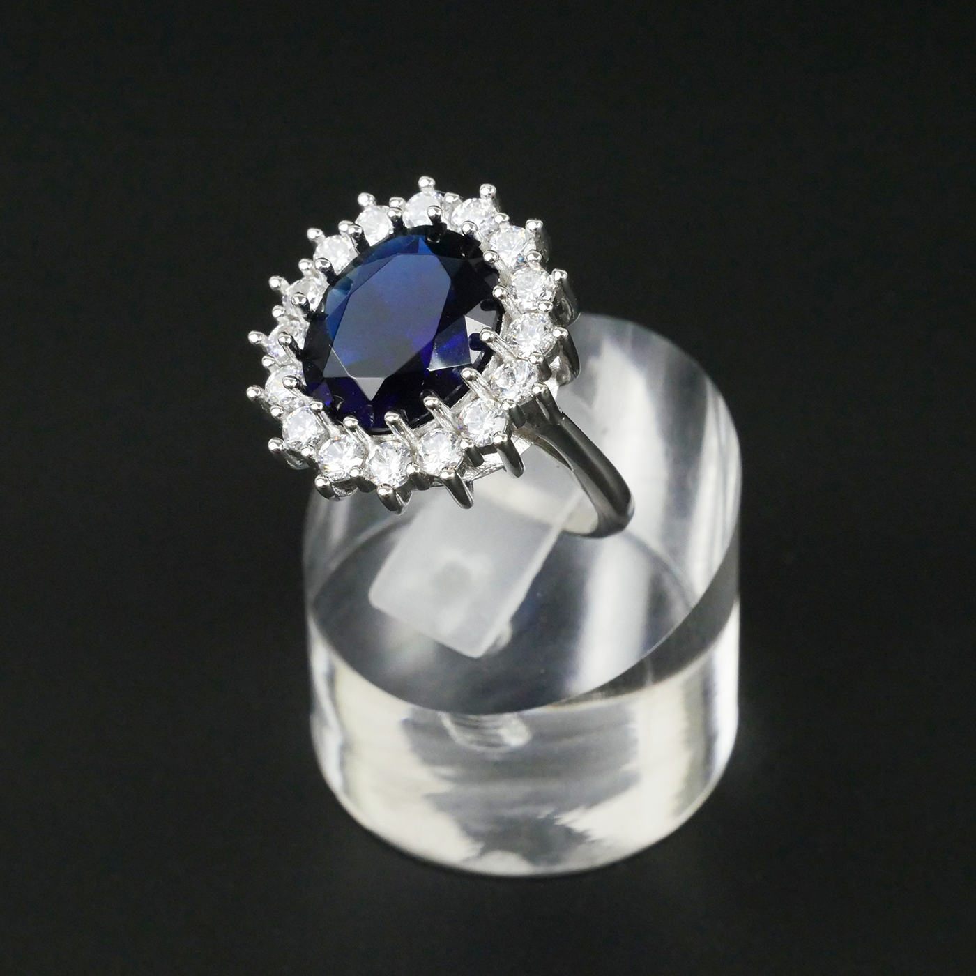 Ring with a blue gemstone and CZ-studded halo on a clear stand against a black background