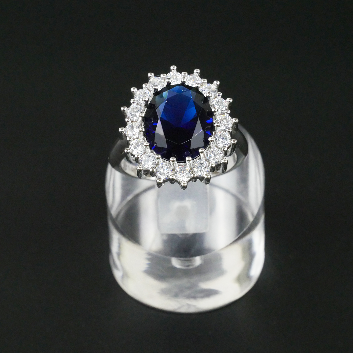 Ring with a blue gemstone and CZ-studded halo on a clear stand against a black background