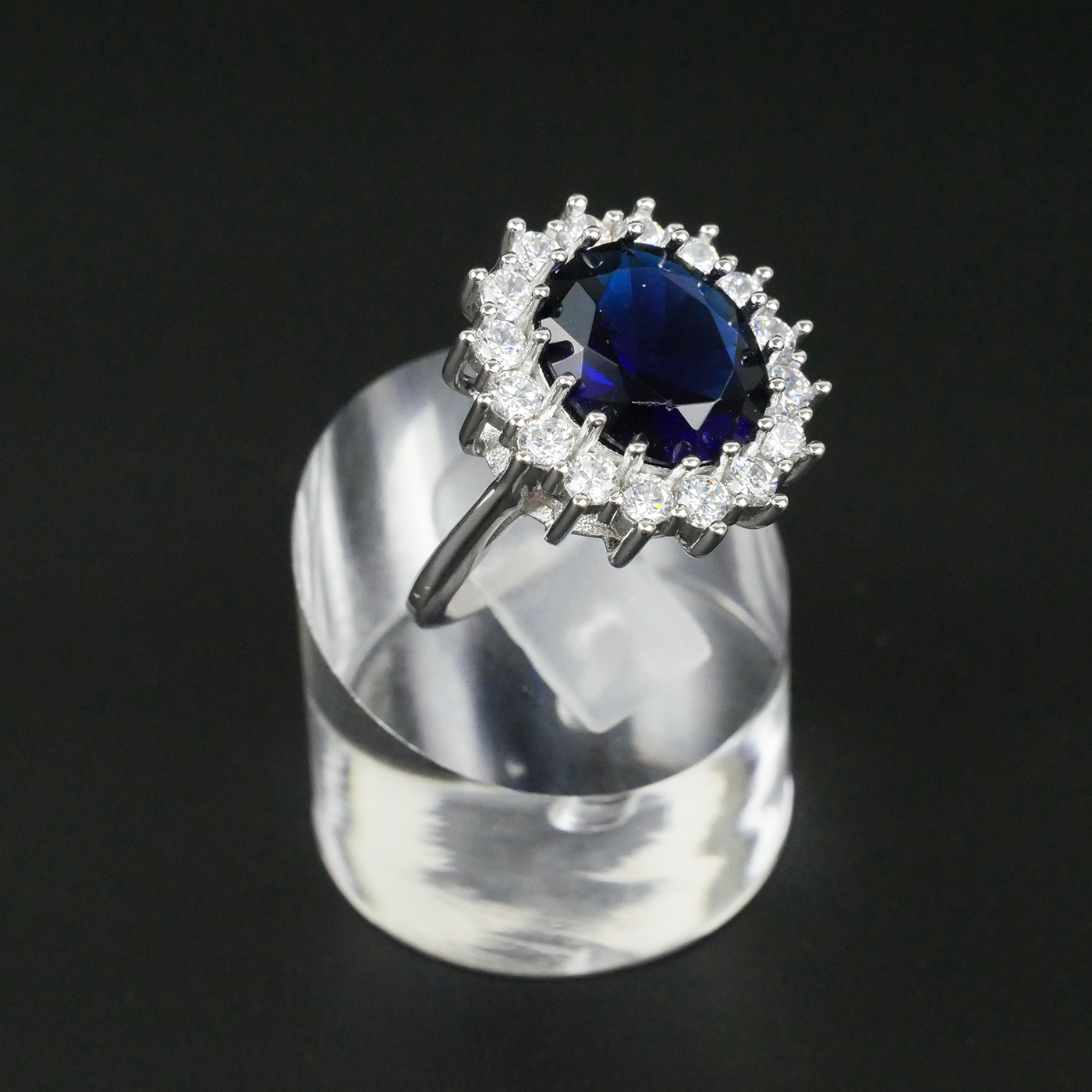 Ring with a blue gemstone and CZ-studded halo on a clear stand against a black background