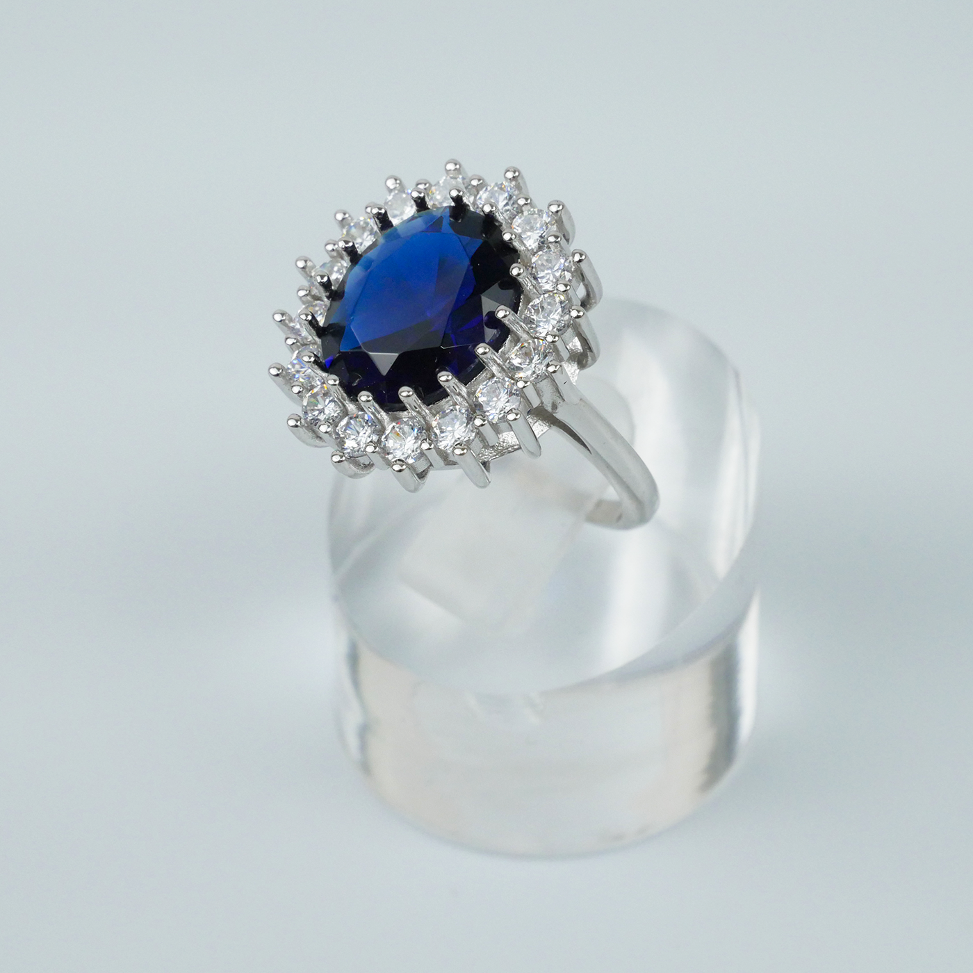 Ring with a blue gemstone and CZ-studded halo on a clear stand against a light-grey background