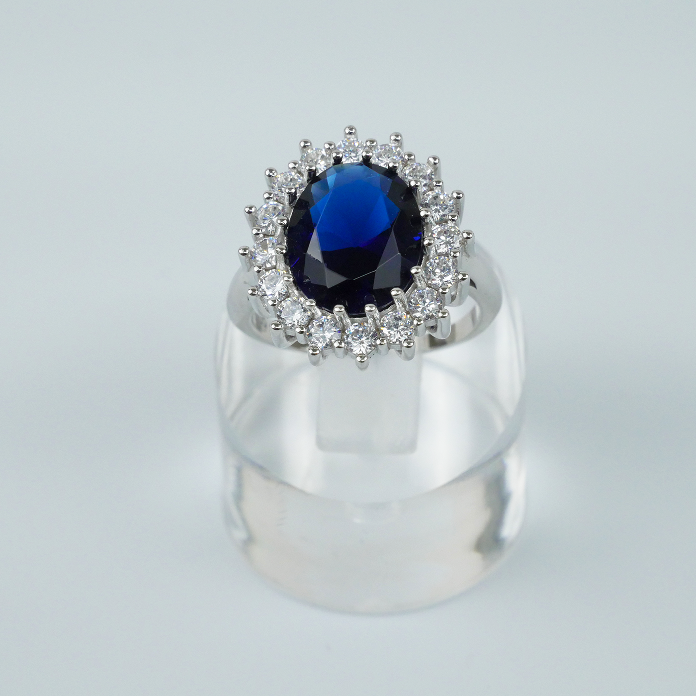 Ring with a blue gemstone and CZ-studded halo on a clear stand against a light-grey background
