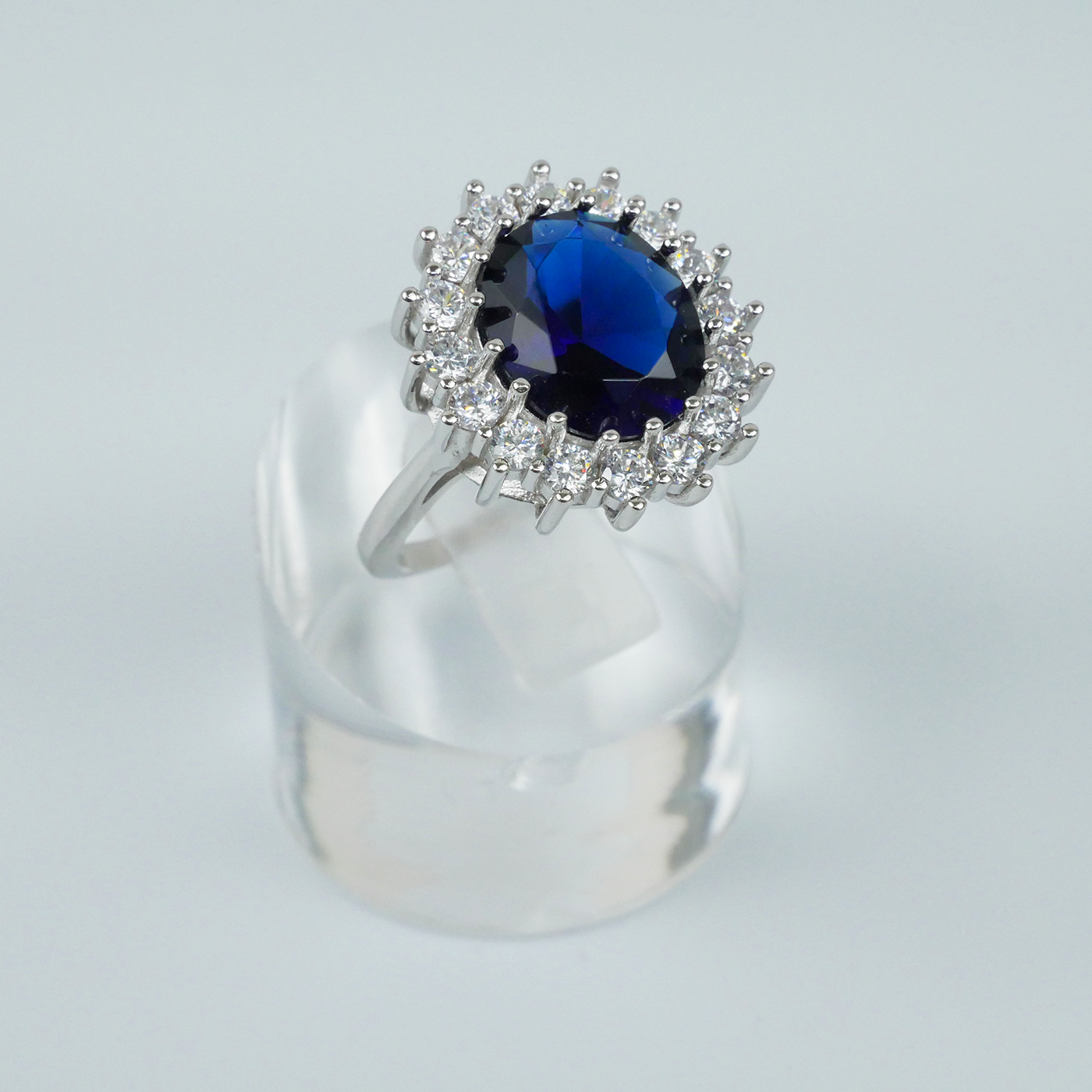 Ring with a blue gemstone and CZ-studded halo on a clear stand against a light-grey background