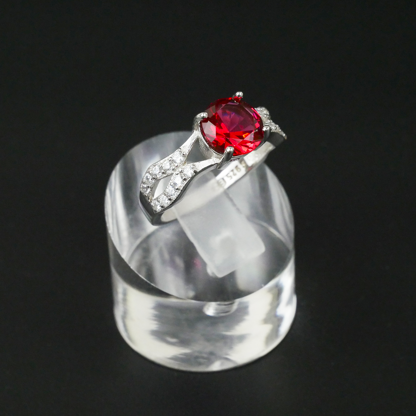Silver ring with a red gemstone on a clear stand against a black background