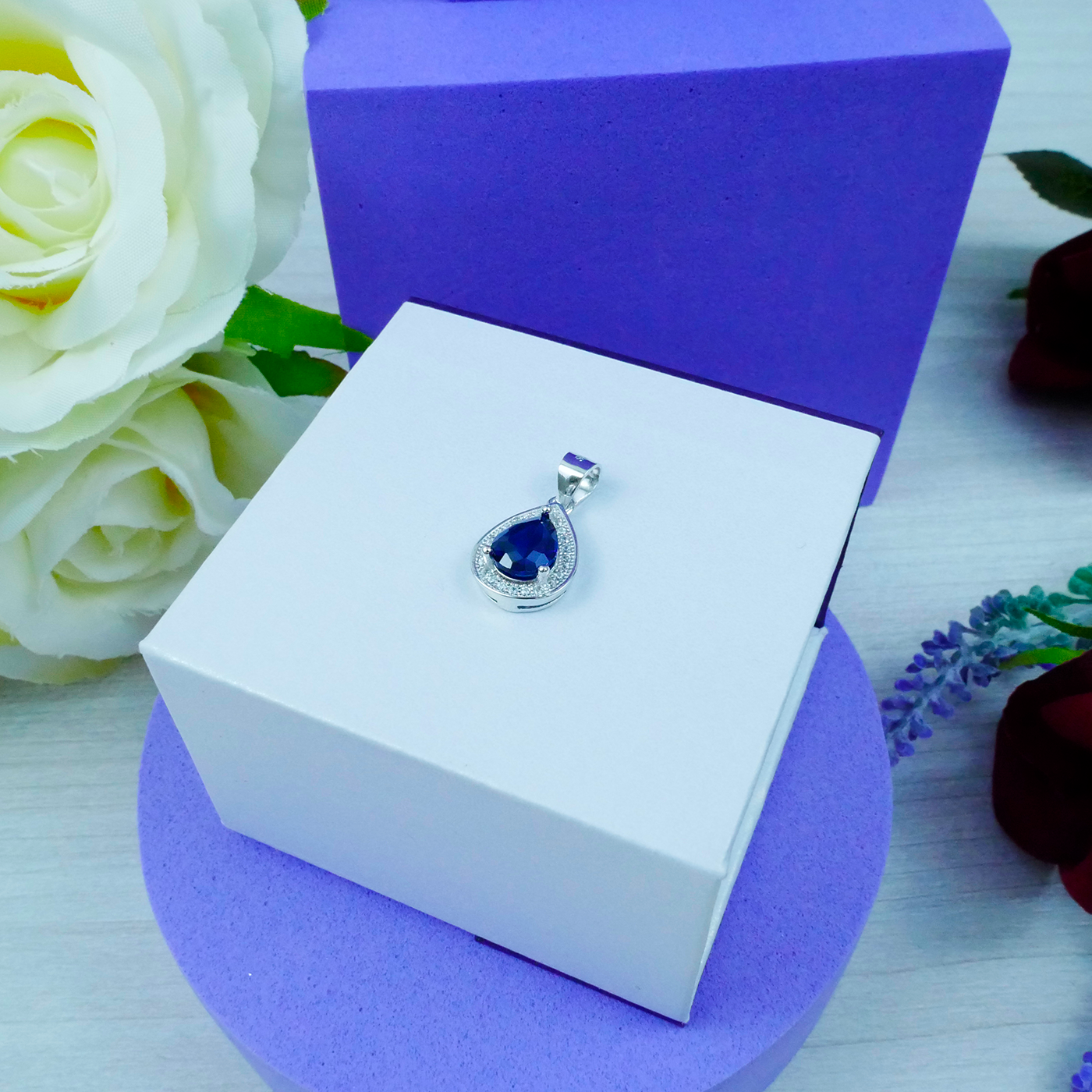 Jewelry pendant with blue gemstone in a white box on a purple base, surrounded by flowers.