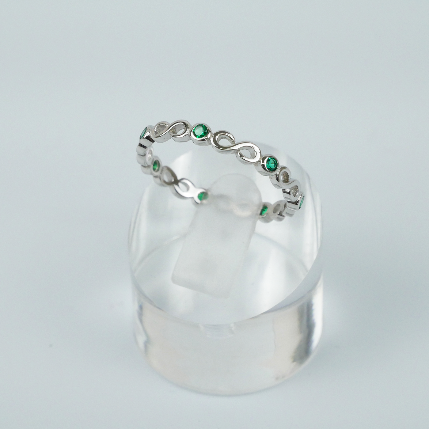 Silver ring with green gemstones on a clear stand against a light gray background