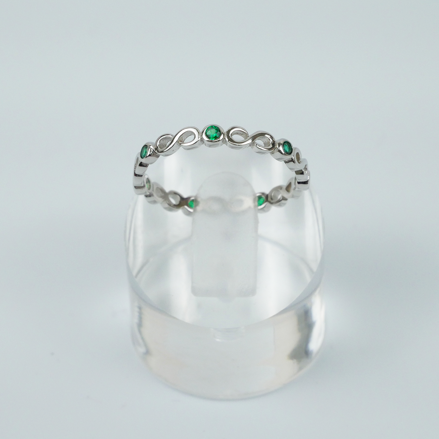 Silver ring with green gemstones on a clear stand against a light gray background