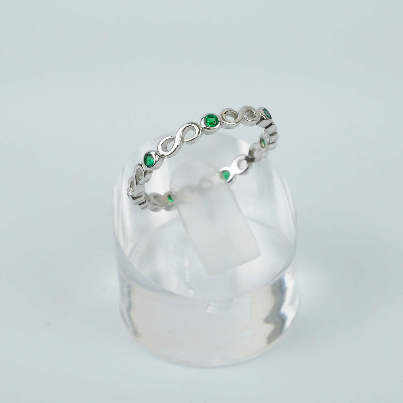 Silver ring with green gemstones on a clear stand against a light gray background