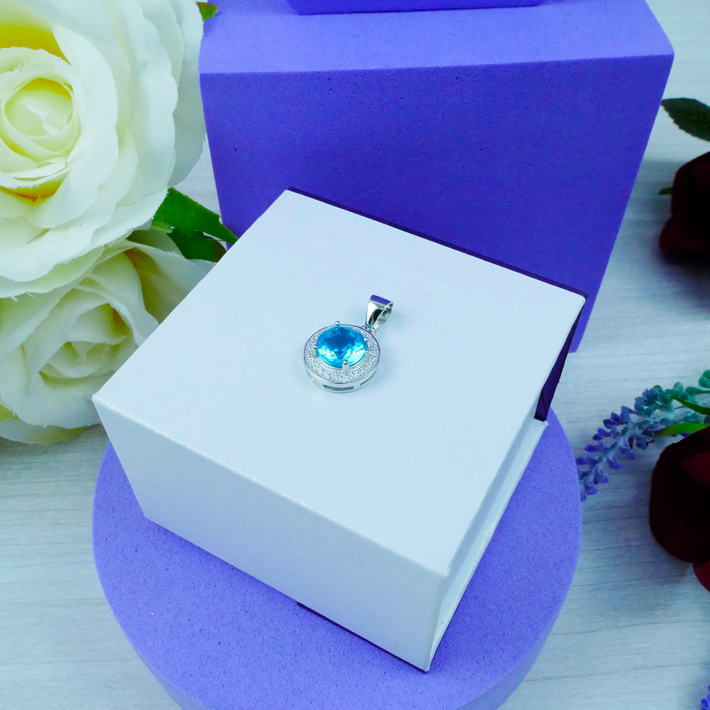 Jewelry pendant with blue gemstone in a white box on a purple stand, surrounded by flowers.