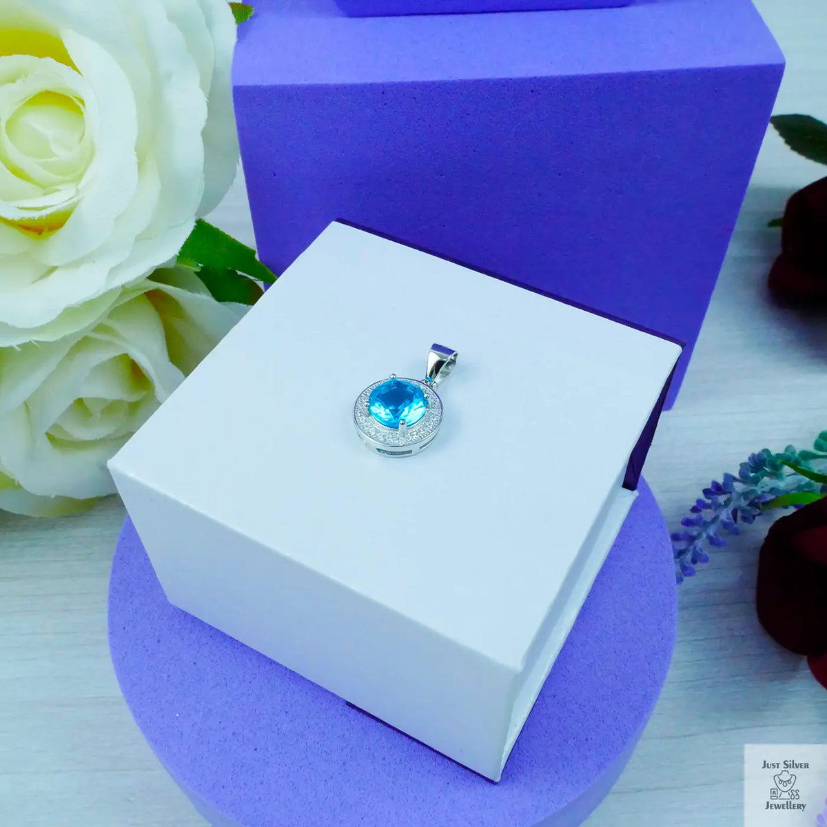 Jewelry pendant with blue gemstone in a white box on a purple stand, surrounded by flowers.