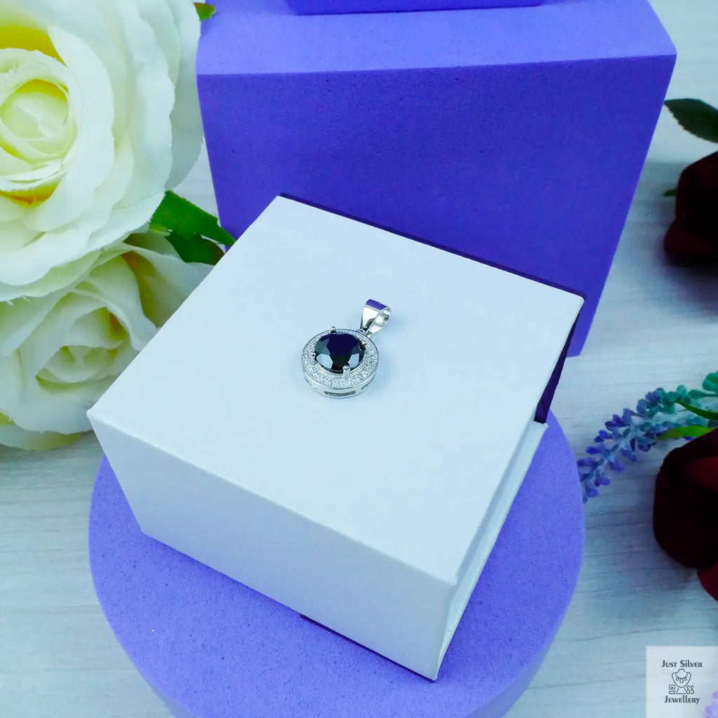 Silver pendant with a blue gemstone on a white box with a purple base, surrounded by flowers.