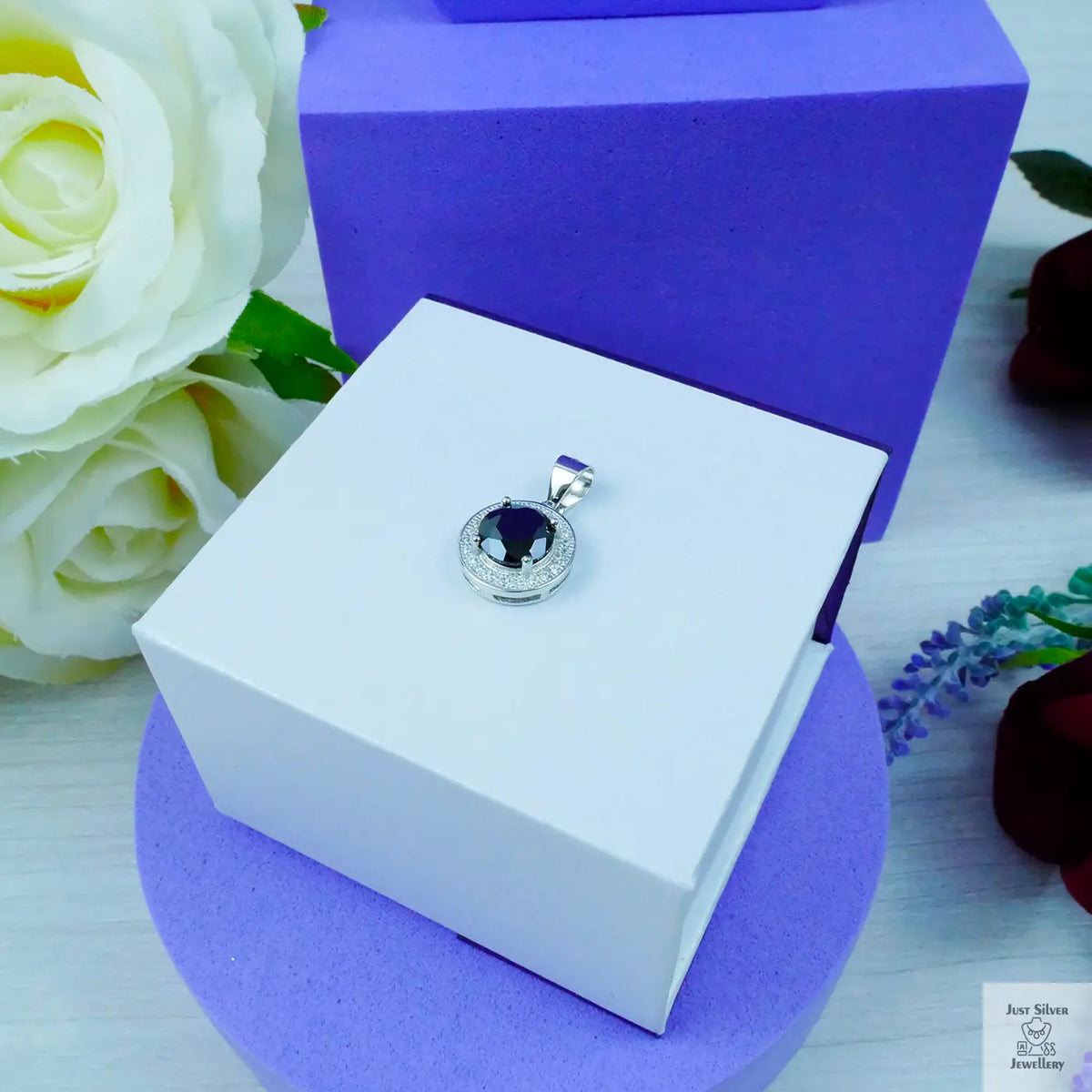 Silver pendant with a blue gemstone on a white box with a purple base, surrounded by flowers.