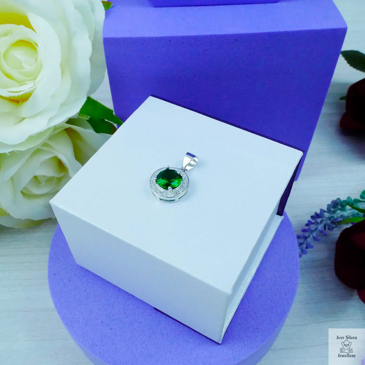 Silver pendant with a green gemstone on a white box with purple base, surrounded by flowers.