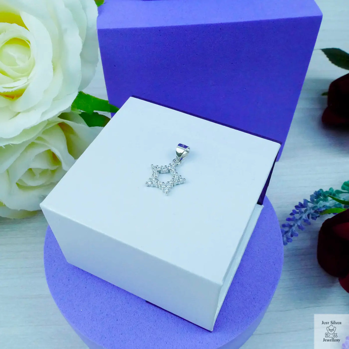 Silver star-shaped pendant on a white box with purple base, surrounded by flowers.