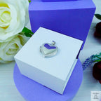 Heart-shaped silver pendant in a white box with purple accents, surrounded by flowers.