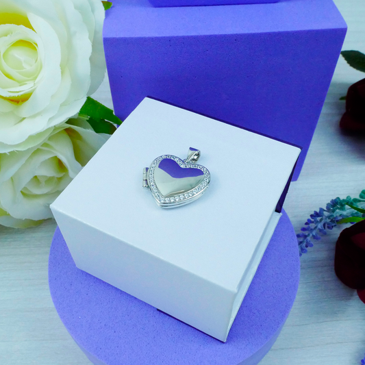 Heart-shaped silver pendant in a white box with purple accents, surrounded by flowers.