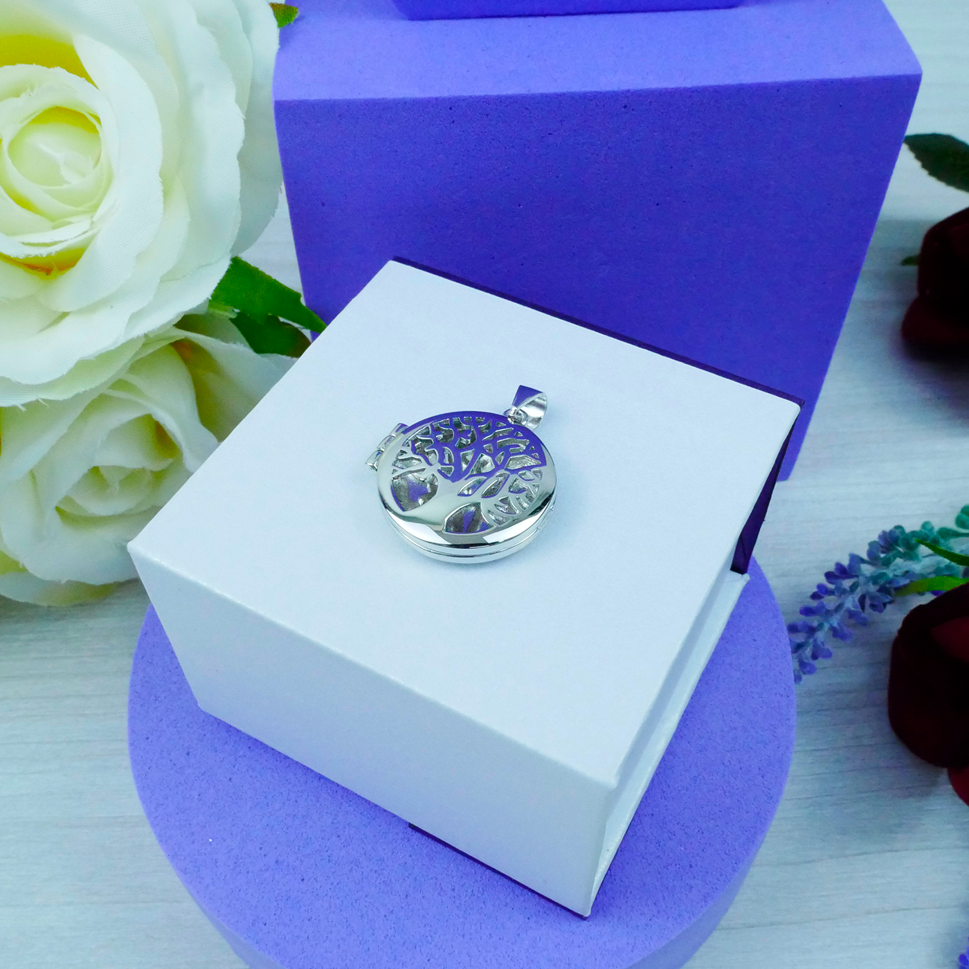 Silver pendant in a white box with purple lid, surrounded by flowers on a light wooden surface.