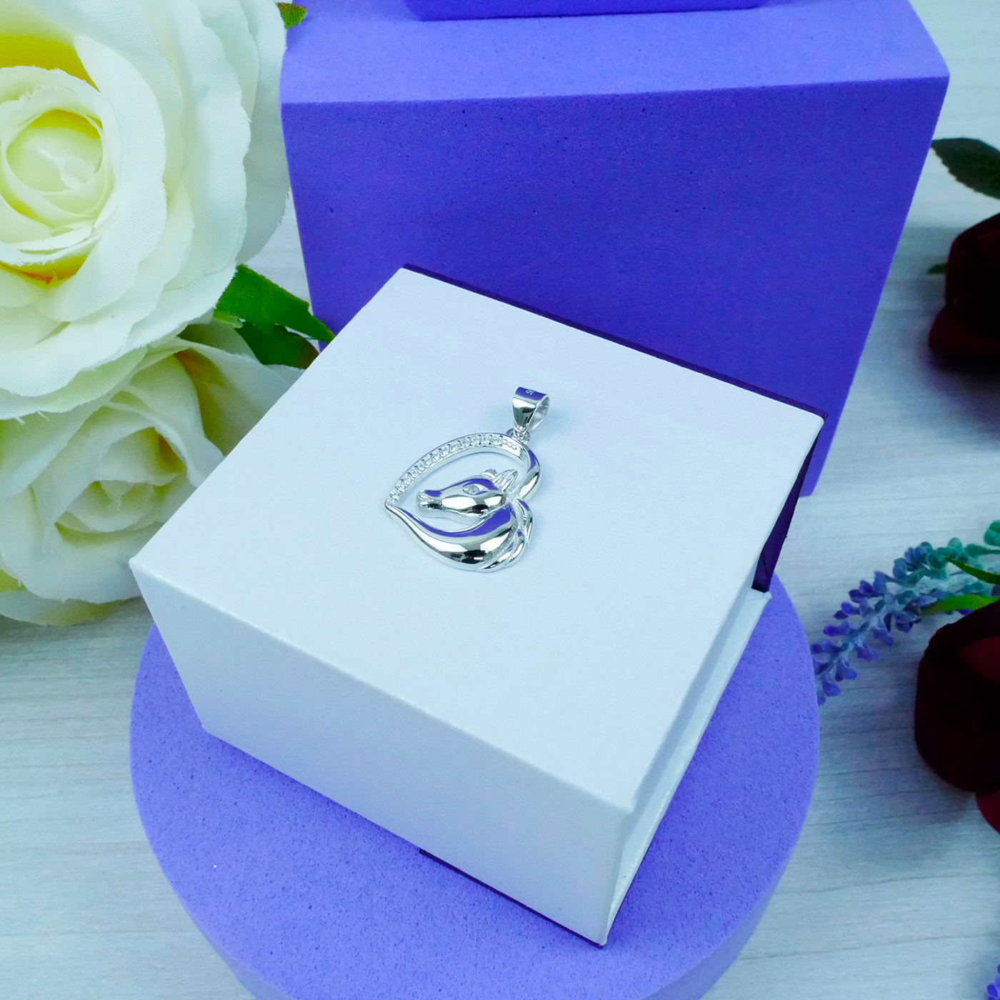 Silver pendant in a white box with purple base, surrounded by flowers